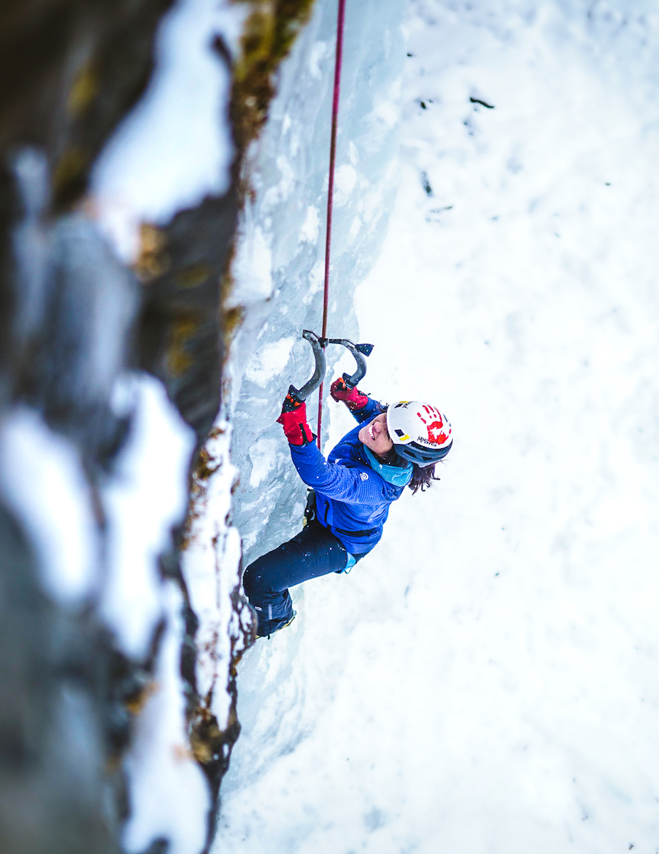 Author Micheli Oliver ice climbing for the first time, with an organization called Inclusive Outdoors, in the shared territories of Blackfeet, Apsaalooke, Confederated Salish and Kootenai, Cheyenne (Tsitsistas and Suhtaio) and Ochethi Sakowin, Hyalite Canyon, Montana. Oliver is wearing the red handprint on her helmet to begin conversations and to raise awareness of MMIWG2S. [Photo] Chris Donovan