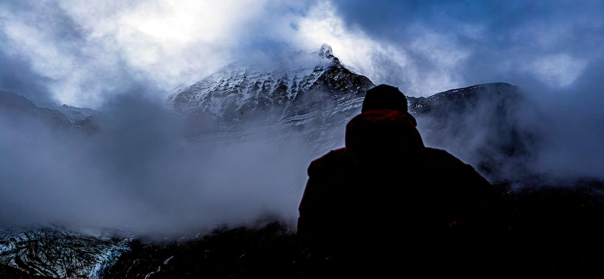 Ethan Berman looking at the Emperor Face from their camp by the river. This was the best view he and Uisdean Hawthorn had before the mist returned. The Texqakallt Nation name for the 3954-meter peak, Yexyexescen, roughly translates to striped rock, a reflection of the multilayered horizontal striations. [Photo] Uisdean Hawthorn