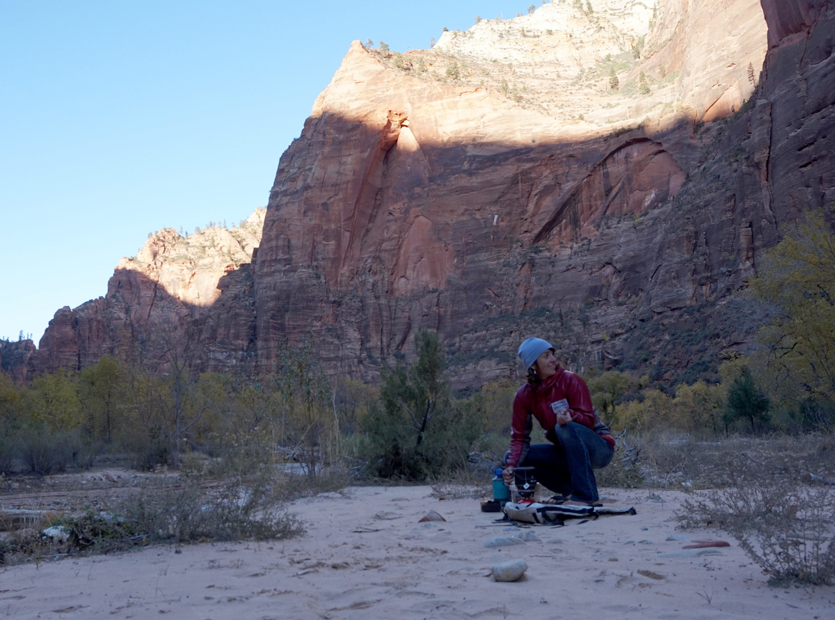 Brett boils water on the banks of the Virgin River with the Leaning Wall in the background. Spaceshot's crack system parallels the left side of the huge dihedral's arete. [Photo] Max Barlerin